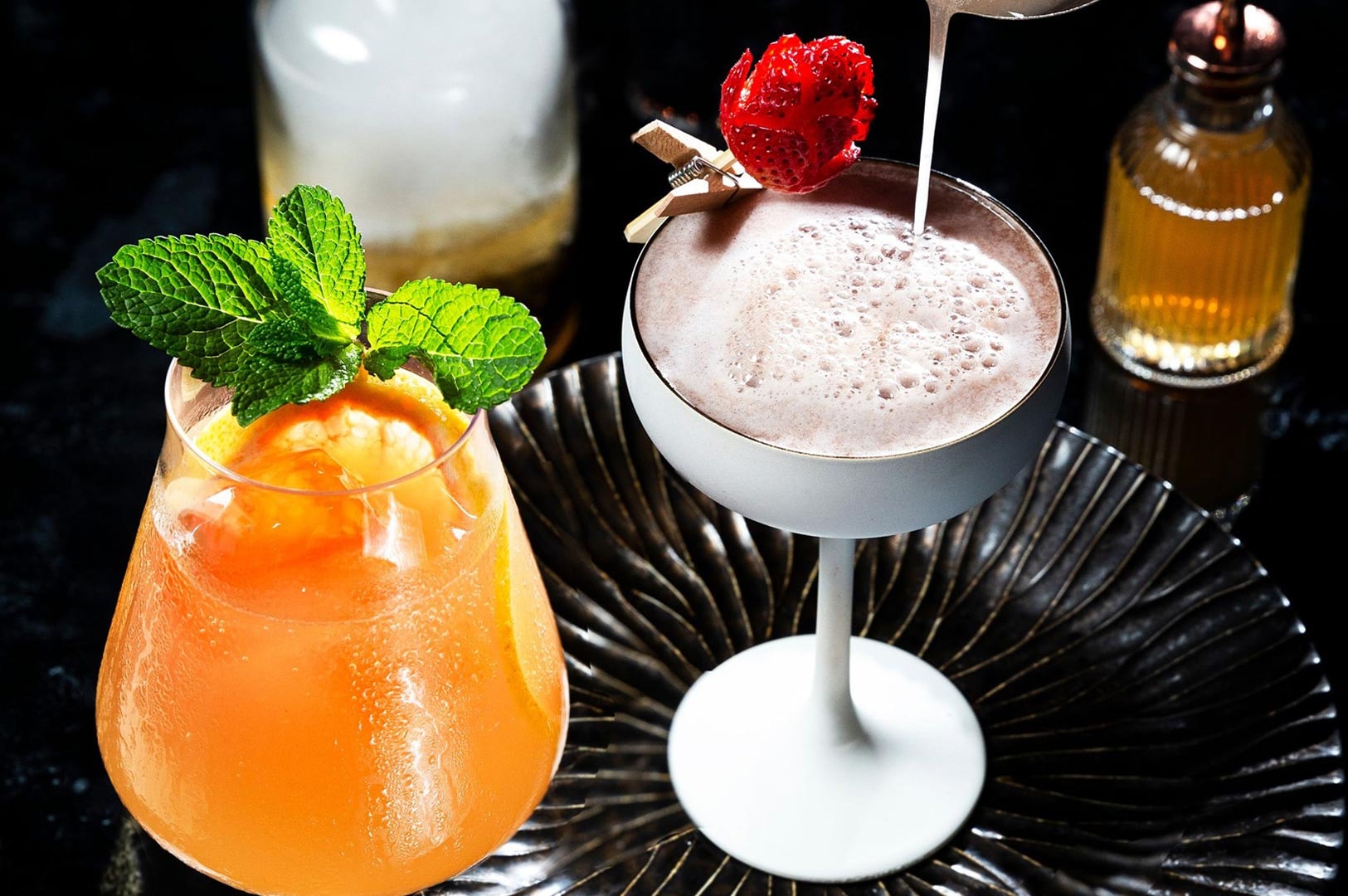 Close-up of two signature craft cocktails; one orange citrus drink with fresh mint and one creamy pink martini with a carved strawberry rose, set against a dark background.