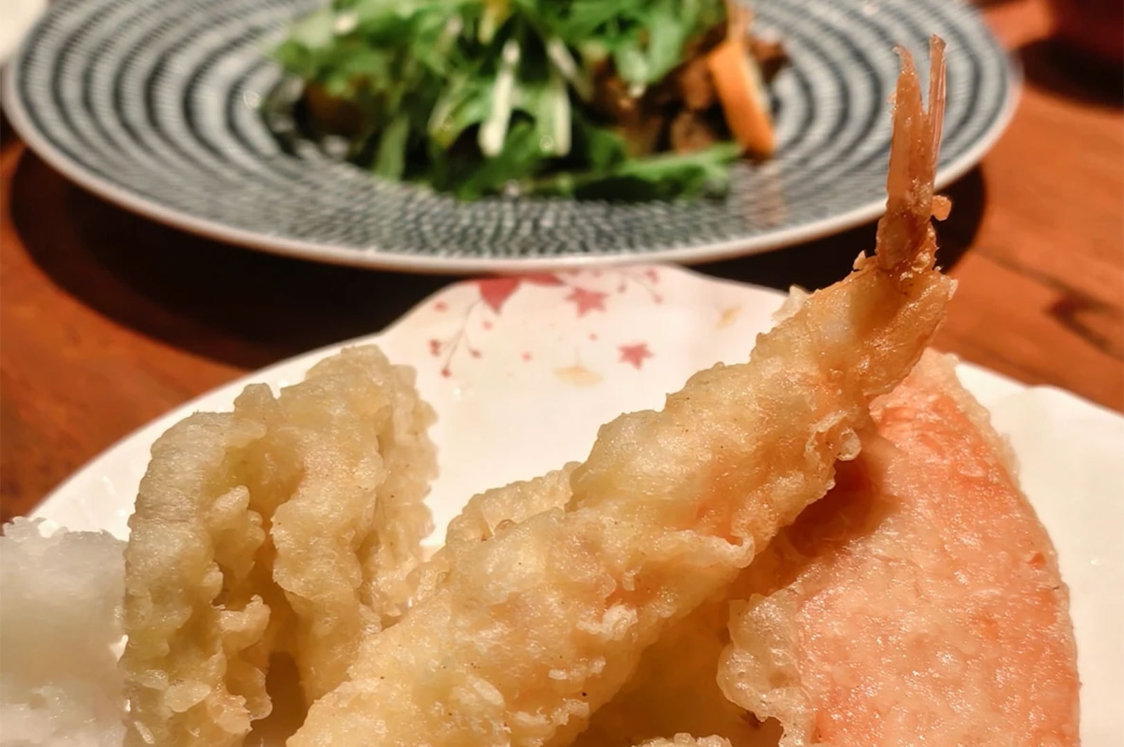 Close-up of golden, crispy shrimp tempura served on a white plate, with a fresh green salad on a blue patterned plate in the background.