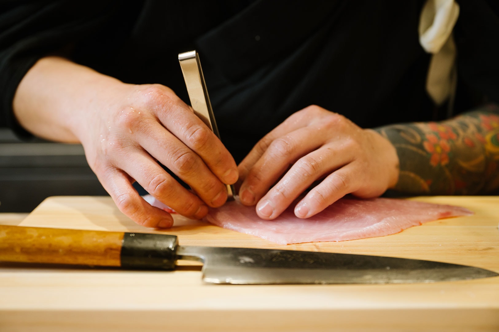 Close-up of a sushi chef's hands using tweezers to remove pin bones from a raw fish fillet next to a traditional Japanese knife.