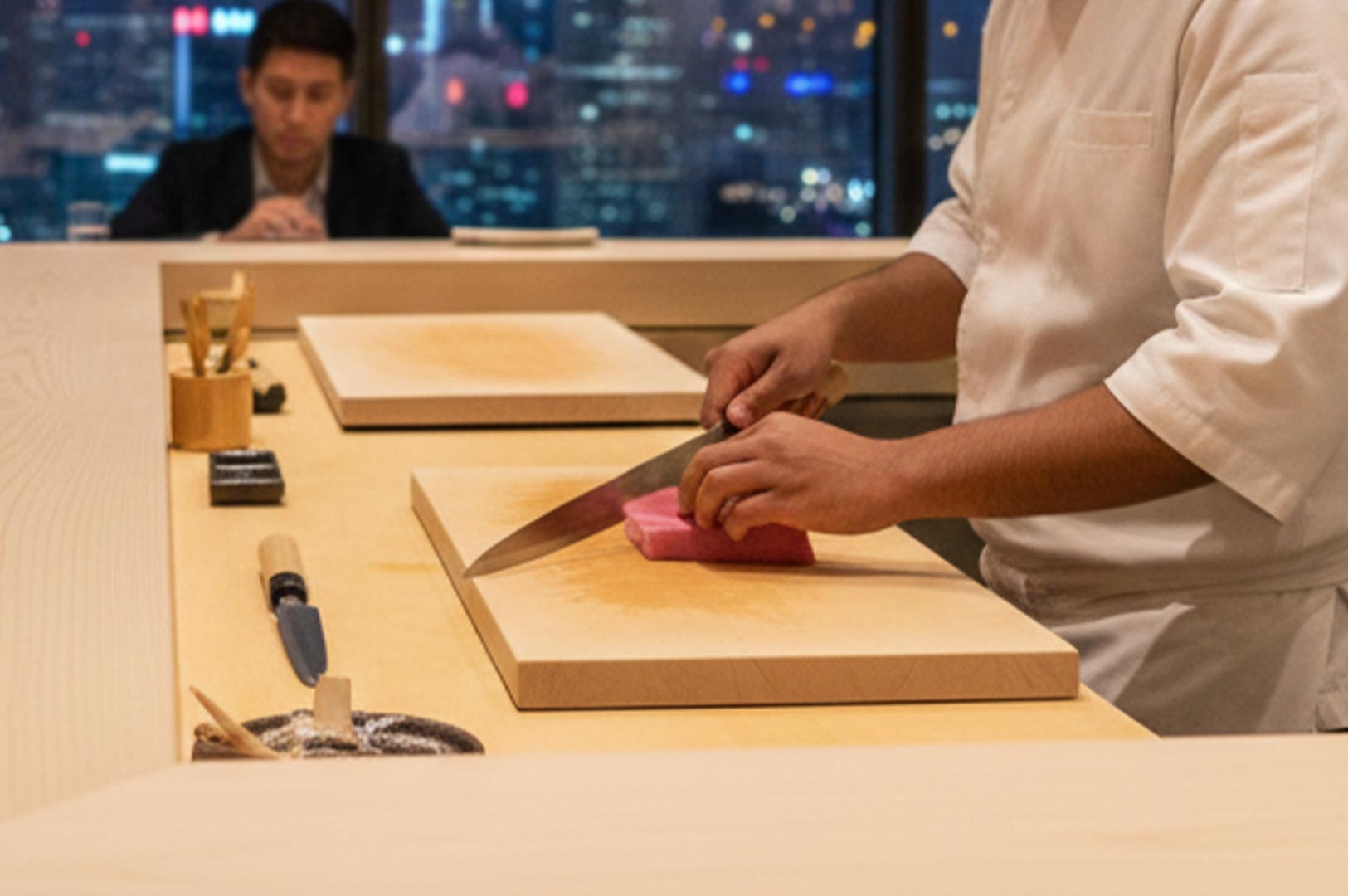 A sushi chef slicing a block of fresh tuna on a wooden board, with a dining customer and blurred city night lights visible in the background.