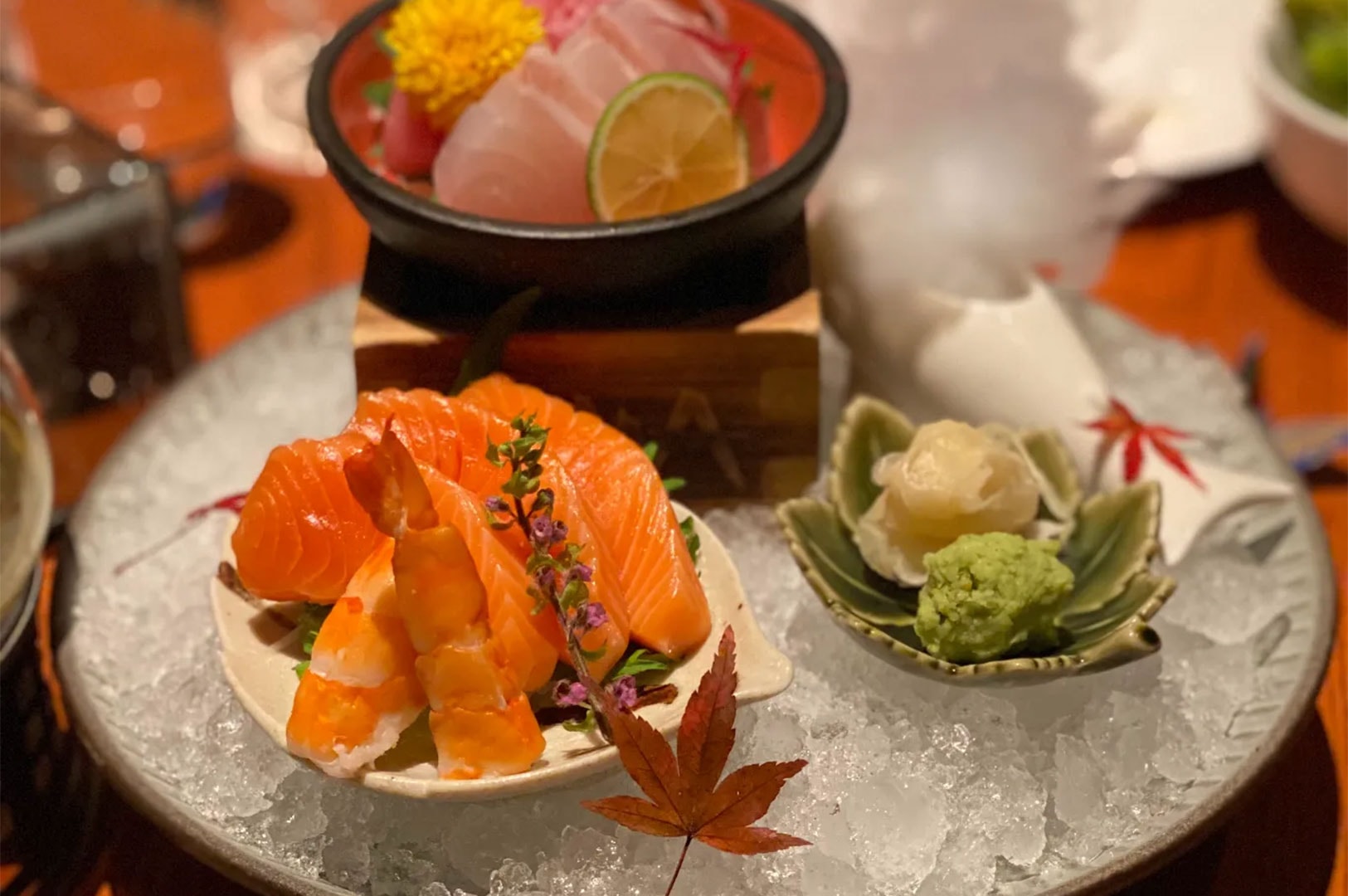 Premium sashimi platter presented on crushed ice, featuring fresh slices of salmon and cooked shrimp in the foreground, with a bowl of white fish sashimi, lime, and yellow flower garnish in the background.