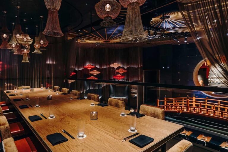 A long wooden communal dining table set for dinner in a dimly lit, upscale Asian fusion restaurant, featuring oversized woven basket pendant lights and a view of a decorative red bridge railing.