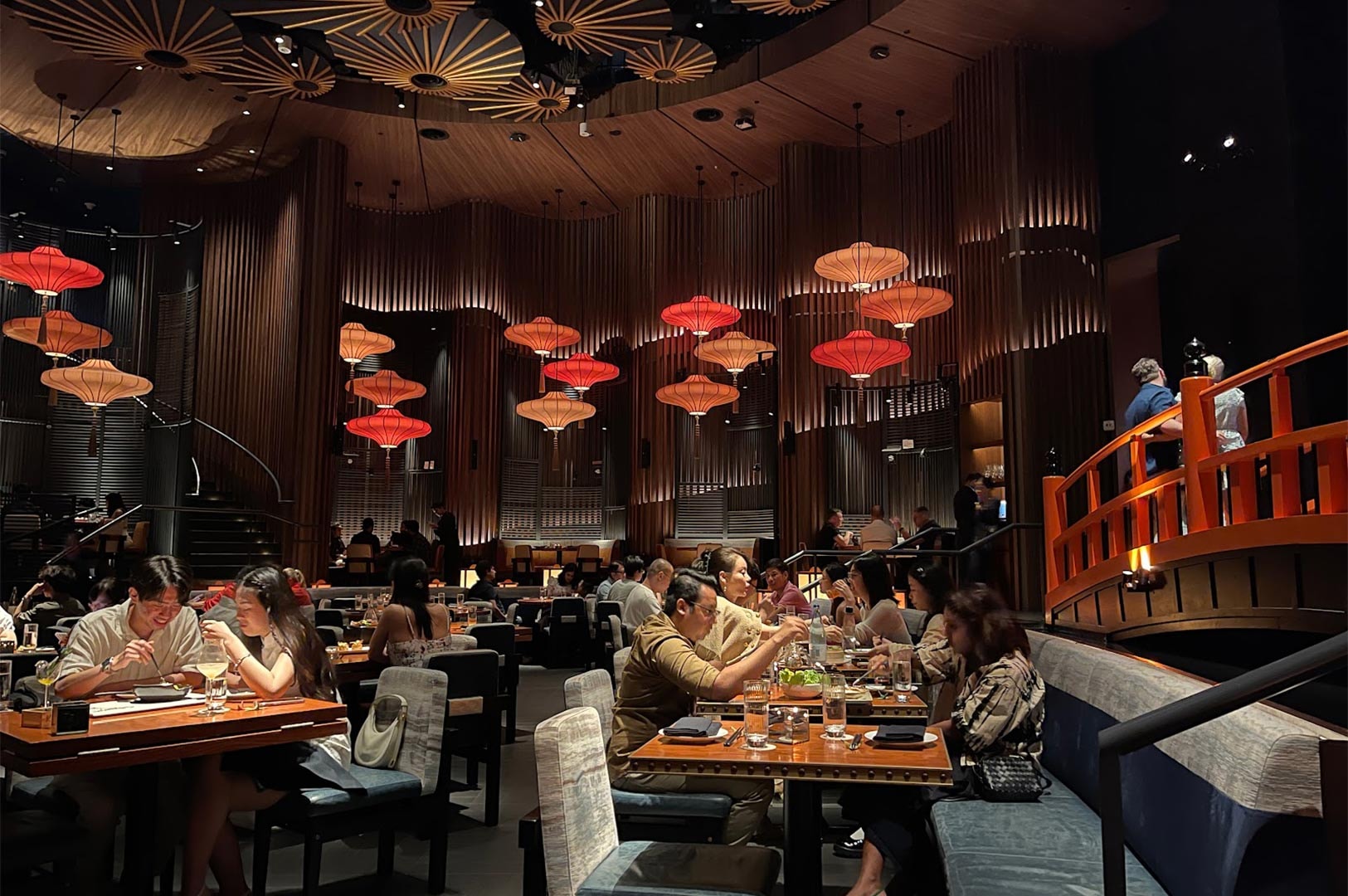 Wide shot of a bustling main dining hall in a high-end restaurant, featuring towering wooden slat walls, floating red lantern lights, and a mezzanine level with orange architectural railings.