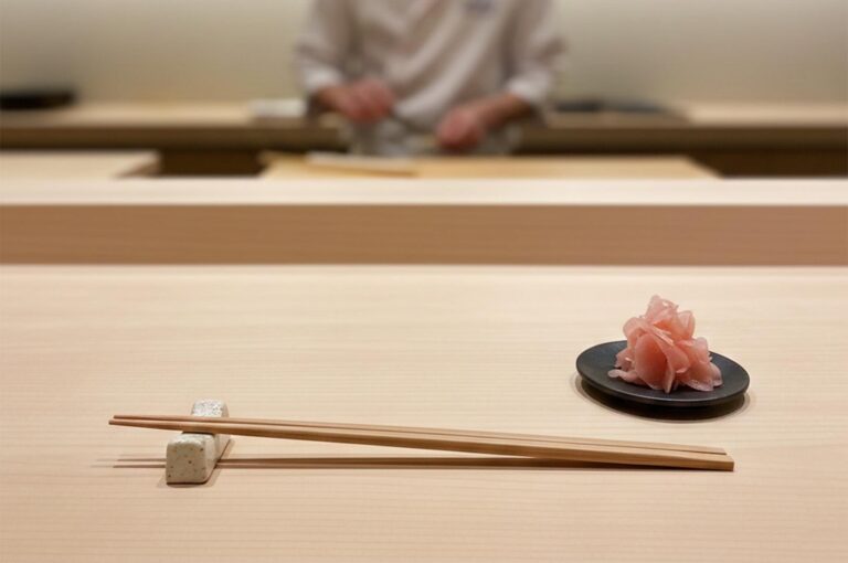 A minimalist wooden sushi counter setting featuring chopsticks on a ceramic rest and a small plate of pickled ginger, with a blurred sushi chef working in the background.