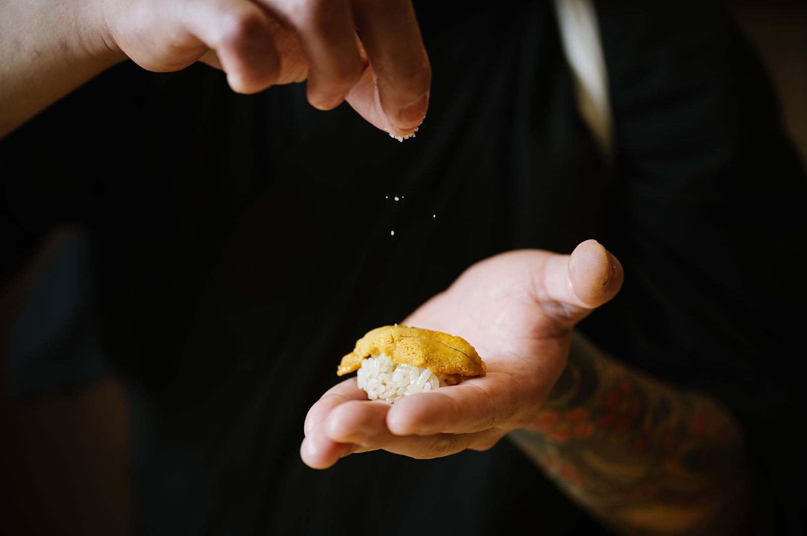 A sushi chef's hands carefully sprinkling seasoning onto a piece of fresh uni (sea urchin) nigiri.