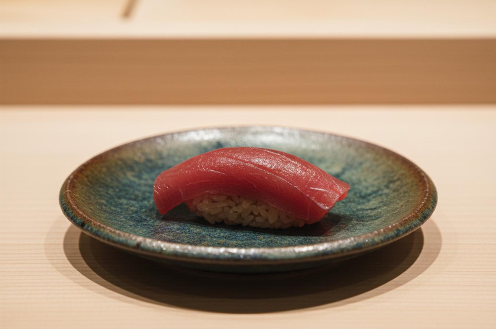 A single piece of premium Maguro (tuna) nigiri sushi served on a handcrafted teal and brown glazed ceramic plate over a light wood counter.
