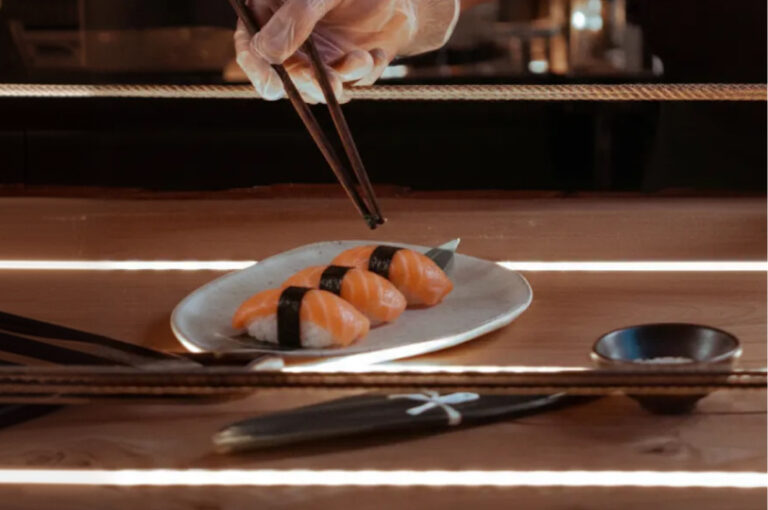 A chef's gloved hand uses chopsticks to garnish three pieces of salmon nigiri resting on a white plate atop a wooden counter.