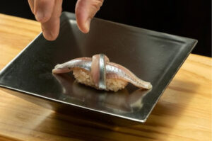 A chef's hand placing a piece of Sayori (Japanese halfbeak) nigiri sushi, featuring a knot of silver-skinned fish over seasoned rice, onto a square black plate.