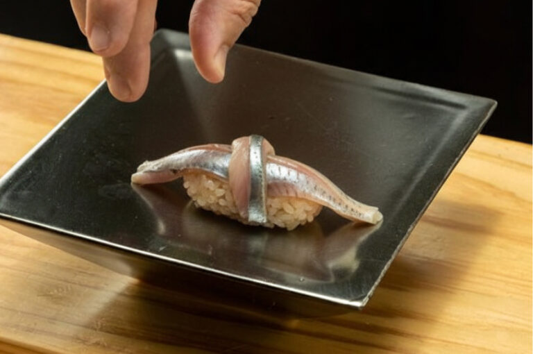 A chef's hand placing a piece of Sayori (Japanese halfbeak) nigiri sushi, featuring a knot of silver-skinned fish over seasoned rice, onto a square black plate.