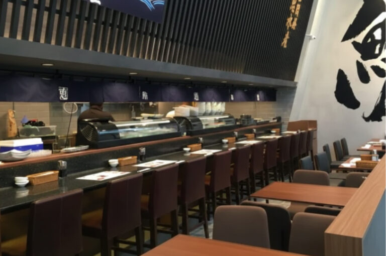 Interior of a Japanese restaurant featuring a long black sushi counter with brown stools and several small wooden tables.