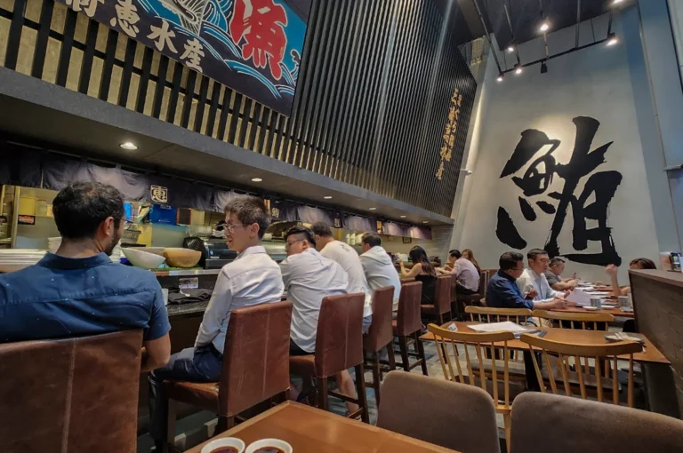 An interior view of the bustling restaurant, capturing diners seated at both the high-backed leather chairs of the sushi bar and wooden dining tables. The high ceiling and minimalist grey walls are accented by a massive black Japanese calligraphy character and warm overhead spotlights, creating a lively urban dining atmosphere.