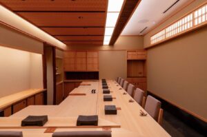 Interior view of a high-end Japanese omakase restaurant featuring a long light-wood counter, minimalist wooden cabinetry, and warm, recessed ambient lighting.