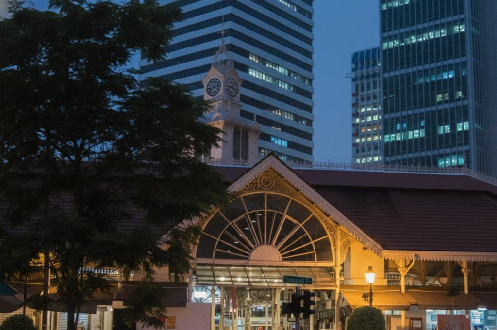 Exterior view of the historic Lau Pa Sat (Telok Ayer Market) in Singapore at night with the clock tower and surrounding skyscrapers.