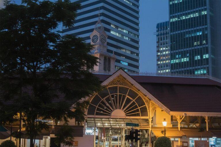Exterior view of the historic Lau Pa Sat (Telok Ayer Market) in Singapore at night with the clock tower and surrounding skyscrapers.