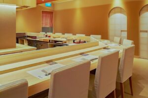 A minimalist, high-end Japanese omakase restaurant interior featuring a light wood sushi counter with elegant white seating.