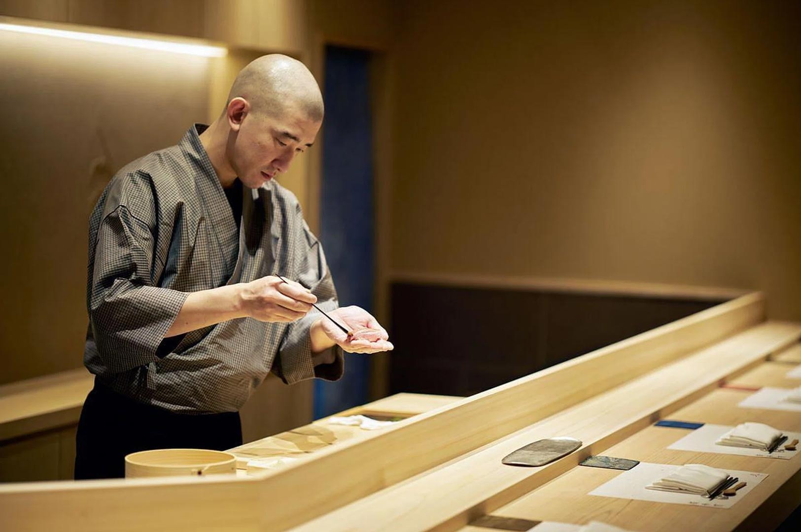 A focused sushi chef in a traditional grey kimono brushing a delicate glaze of nikiri shoyu onto a piece of sushi at an omakase counter.