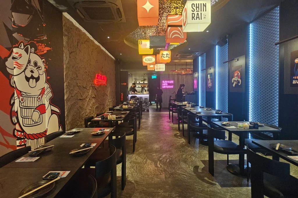 A wide-angle view of a long, narrow Japanese dining room with an edgy, urban aesthetic. The left wall features a large, playful illustration of a white dog in samurai armor next to a section of craggy, brown stone wall. Hanging overhead is a series of illuminated cube lanterns, some featuring the brand name "SHINRAI" and others with Japanese characters. Rows of dark wooden tables are set with black plates and chopsticks. Blue vertical light strips and framed anime-style art line the right wall. In the distant background, staff members are visible working at a lit bar area under a pink neon sign.