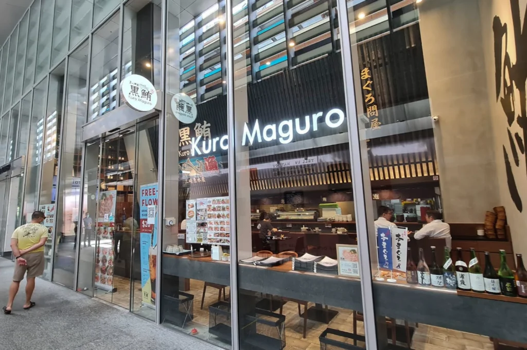 A wide-angle exterior view of the Kuro Maguro restaurant, showcasing its modern glass facade and tall vertical pillars. Through the large windows, customers can be seen seated at a long wooden counter and dining tables, while several sake bottles are lined up on an outdoor ledge.