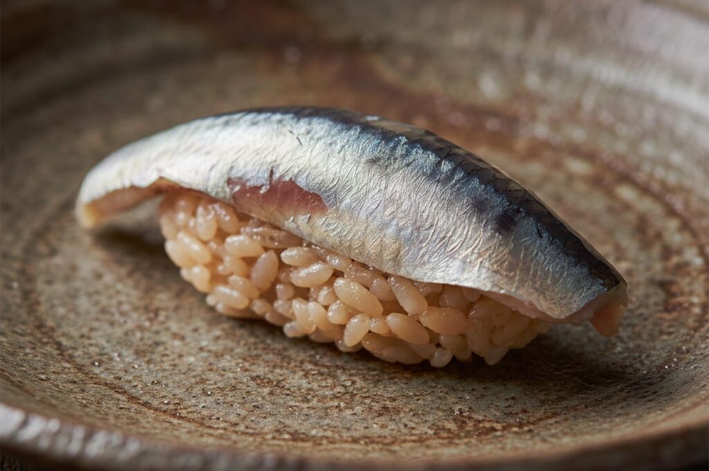 A single piece of Iwashi (sardine) nigiri sushi featuring a shimmering silver skin fillet over seasoned brown sushi rice, served on a rustic ceramic plate.