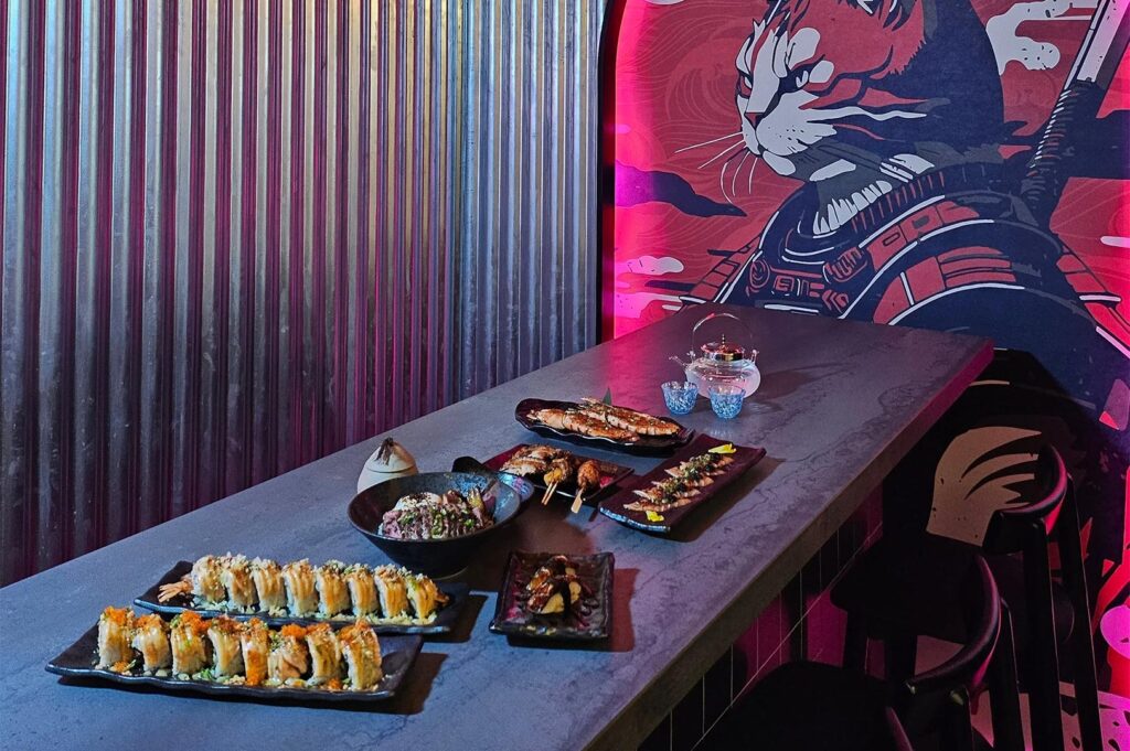 A variety of sushi rolls and Japanese appetizers served on a dark stone table in a modern restaurant featuring a neon samurai cat mural.