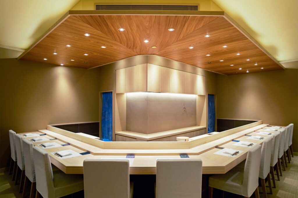 A wide-angle view of a contemporary sushi bar featuring a geometric wooden ceiling with recessed lighting and a wraparound counter.