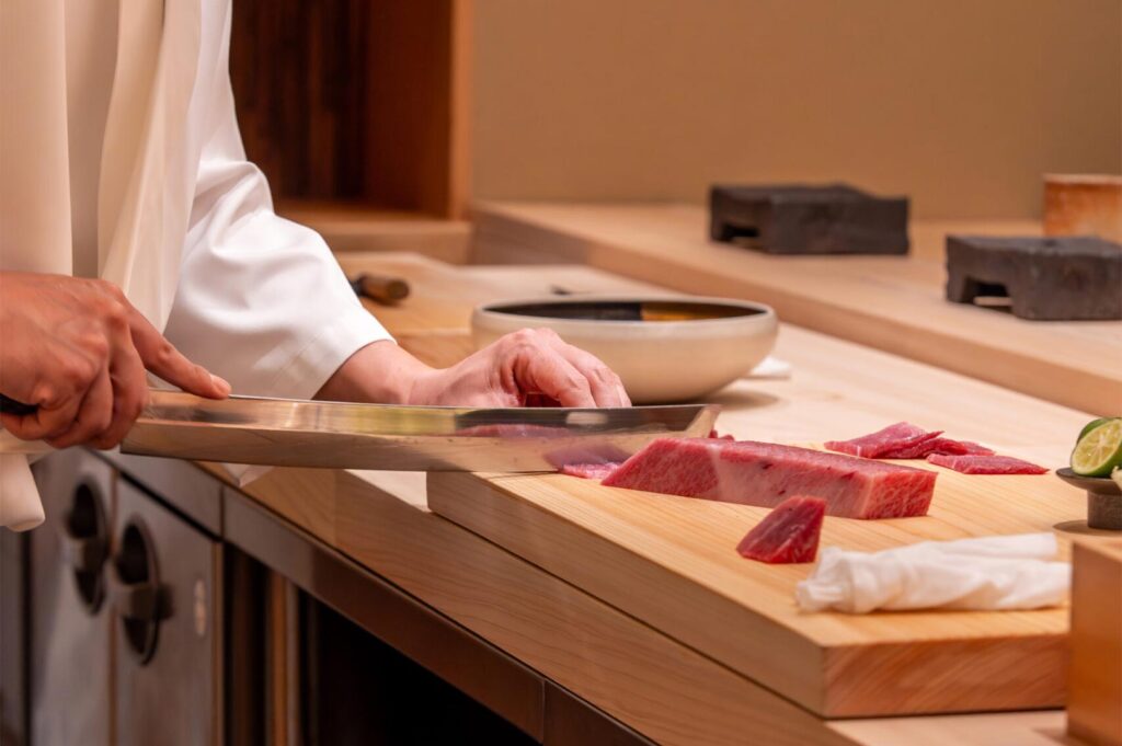 Close-up of a professional sushi chef using a traditional yanagiba knife to precision-cut a marbled piece of fatty tuna (otoro) on a wooden cutting board.