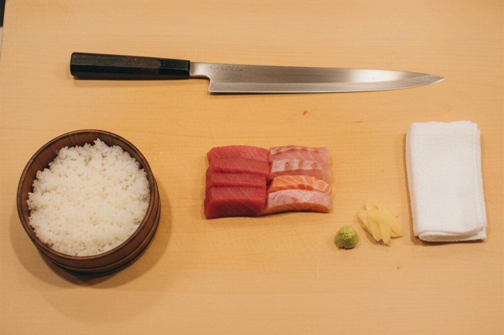A top-down view of sushi preparation components including a Japanese yanagiba knife, sliced tuna and salmon sashimi, a bowl of seasoned rice, wasabi, ginger, and a white hand towel on a light wood cutting board.