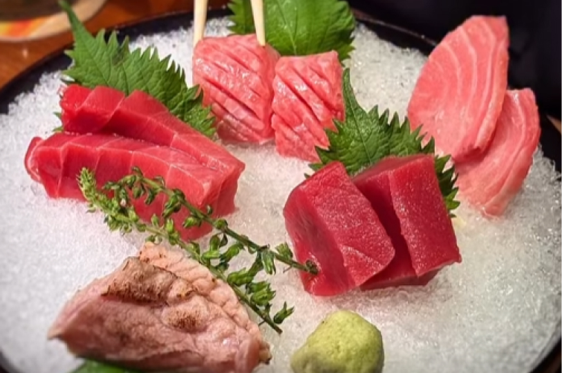 A close-up of a sashimi platter with various cuts of fresh tuna and a seared fish slice served over crushed ice with shiso leaves and wasabi.