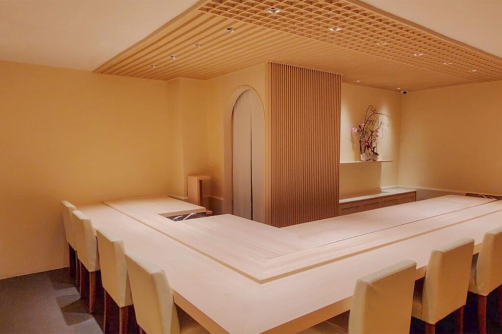 An empty, minimalist Omakase dining room featuring a clean light wood counter, beige chairs, and traditional Japanese slatted wood ceiling.