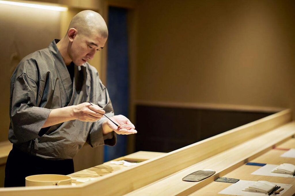 A professional sushi chef in traditional attire meticulously applying a glaze to a piece of fresh nigiri at a wooden counter.