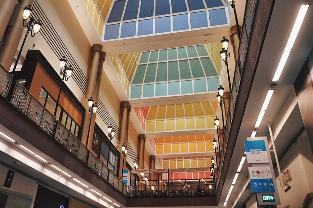 Low-angle architectural shot of a modern multi-level shopping mall atrium featuring a vibrant, multi-colored glass pyramid skylight and classic street-lamp style interior lighting.