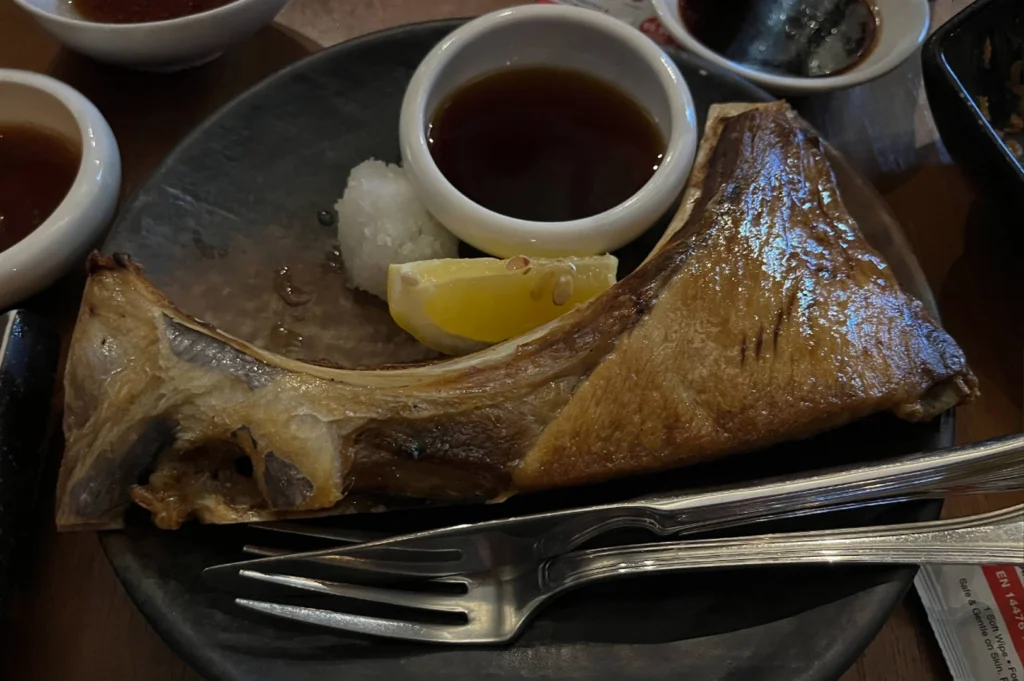 A close-up shot of a large, grilled fish collar served on a dark, rustic plate. The fish skin is a glistening golden brown, accompanied by a small pile of grated daikon radish, a lemon wedge, and a small white bowl of dark dipping sauce, with metal chopsticks and a fork resting in the foreground.