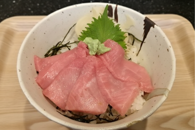 A tuna donburi bowl featuring thick slices of pink tuna sashimi over rice, garnished with a shiso leaf and a dollop of wasabi.