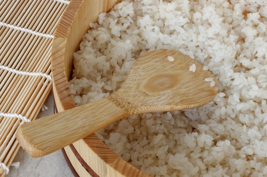 Close-up of seasoned Japanese sushi rice (shari) in a traditional wooden hangiri mixing tub with a bamboo rice paddle and rolling mat nearby.
