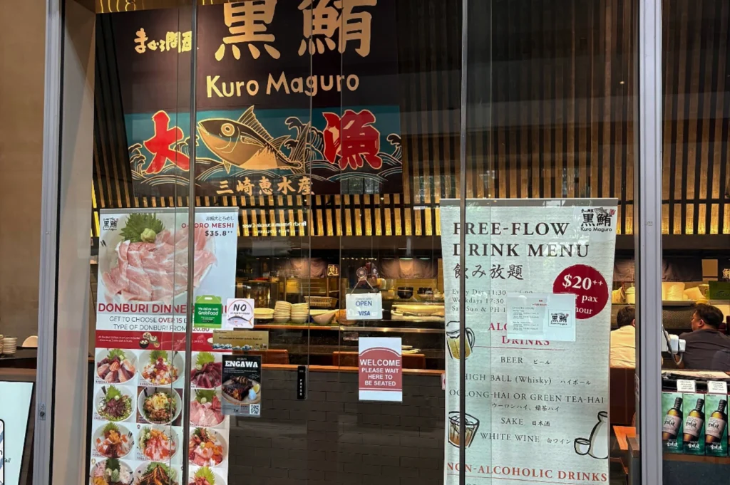 A direct view of the restaurant's glass entrance, cluttered with various promotional posters and signage. A large banner at the top features a blue tuna illustration, while posters on the glass advertise a "Donburi Dinner" with various bowl options and a "$20++ Free-Flow Drink Menu" featuring beer, highballs, and sake.