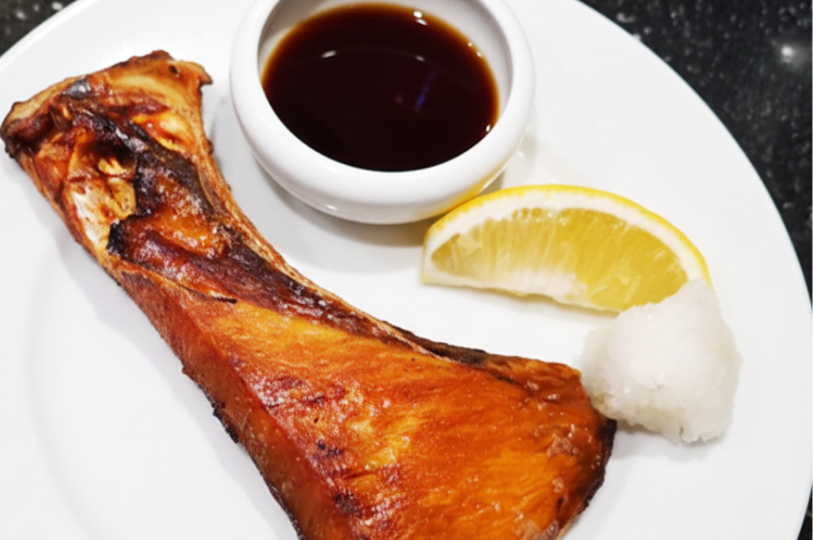 A grilled fish collar served on a white plate with a side of soy sauce, a lemon wedge, and grated daikon radish.