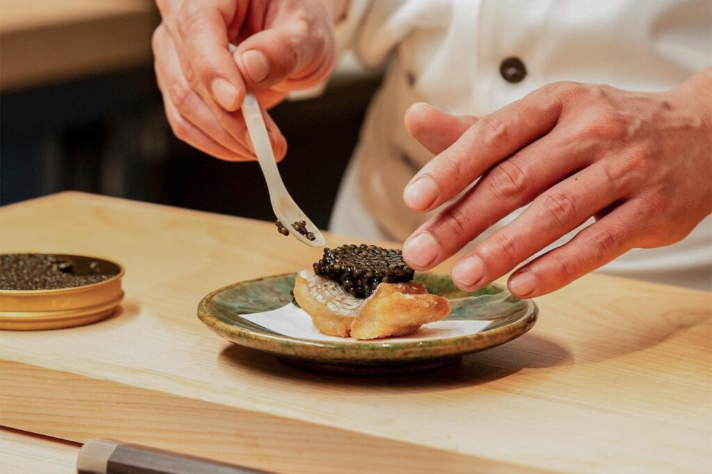 A chef carefully garnishing a piece of seared fish with a generous spoonful of black sturgeon caviar using a mother-of-pearl spoon on a ceramic plate.