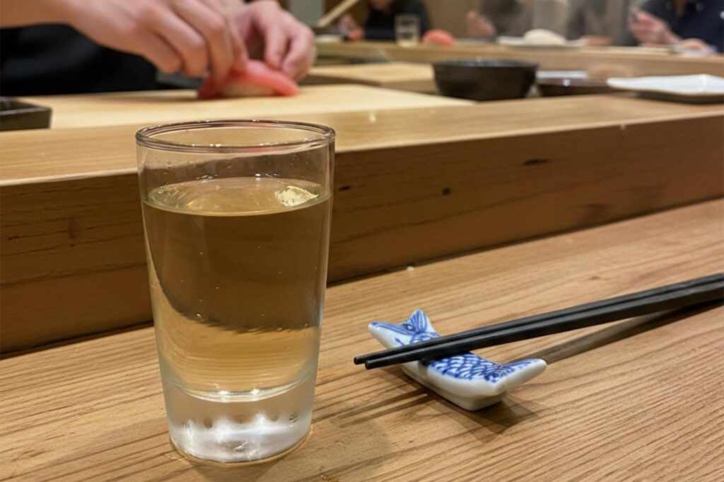 Close-up of a sushi chef’s hands preparing fresh tuna nigiri at a wooden sushi counter next to a glass of tea and chopsticks.