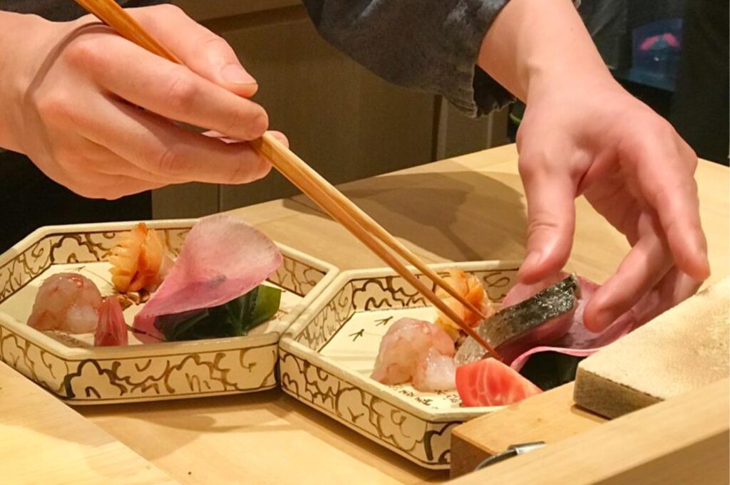 Close-up of a chef using long wooden plating chopsticks to carefully arrange sashimi and garnishes on an ornate ceramic plate.
