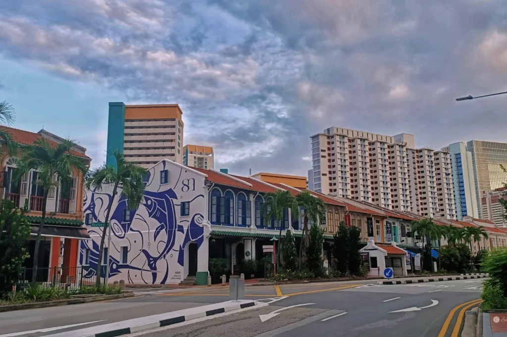 A street-level landscape of Tanjong Pagar, featuring a row of traditional colorful shophouses under a cloudy evening sky. One prominent white building on the corner is decorated with a large blue mural of a fish, while modern high-rise apartments and office buildings loom in the background across the asphalt road.