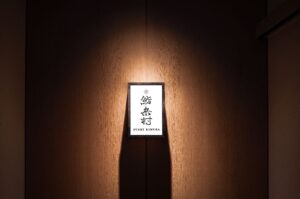 Japanese sushi restaurant signboard “Sushi Kimura” framed on wood, highlighted by overhead light.