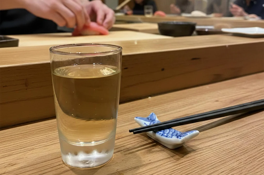 Close-up of a sushi chef's hands preparing fresh tuna nigiri at a wooden sushi counter next to a glass of tea and chopsticks.