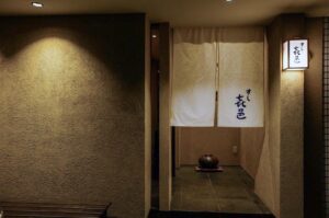 The minimalist exterior entrance of Sushi Nanairo, featuring a traditional white noren curtain with Japanese calligraphy and a warm glowing lantern against a textured clay wall.