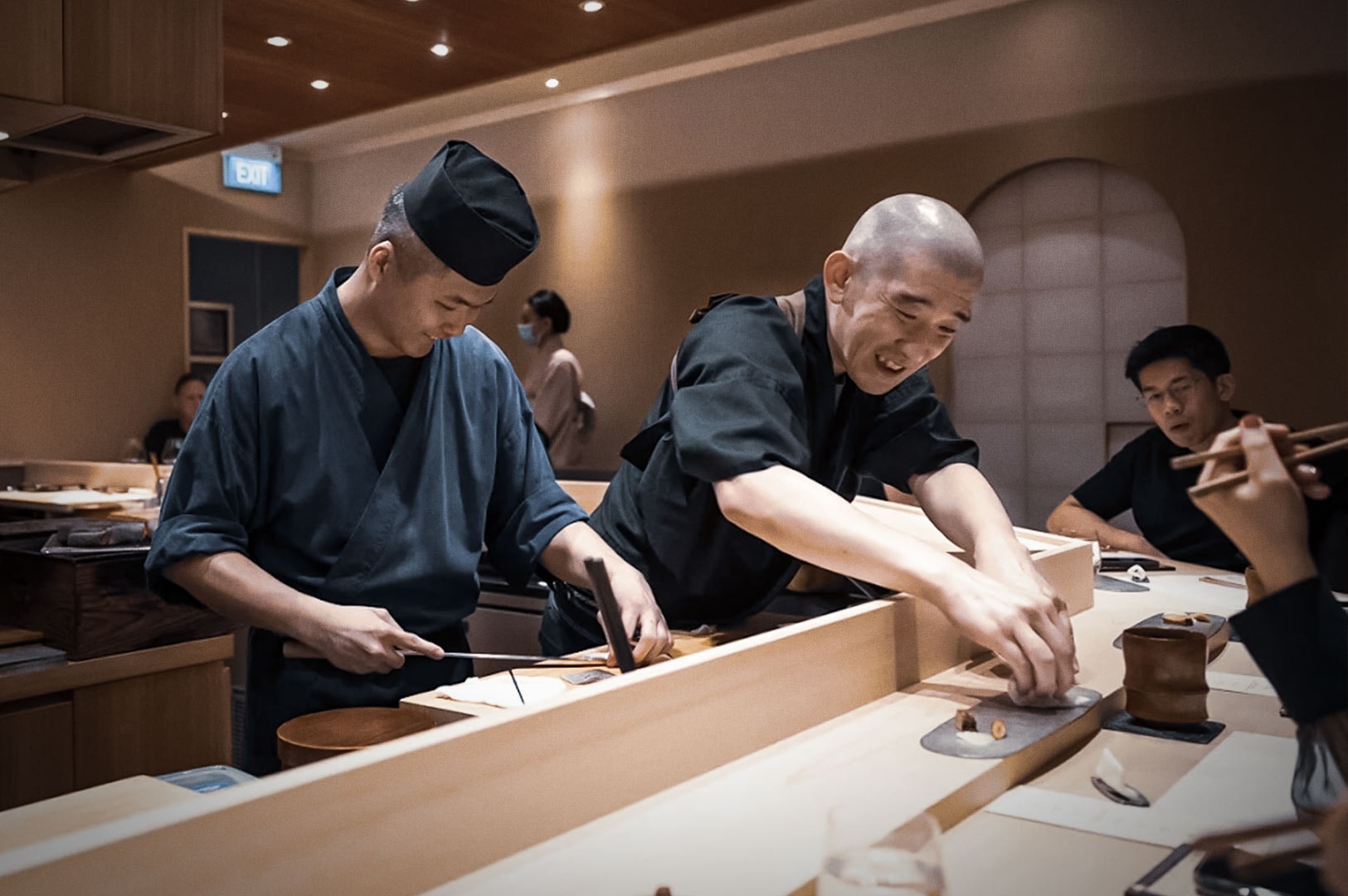 Japanese omakase restaurant scene with sushi chefs in traditional uniforms preparing and serving fresh sushi at wooden counter.