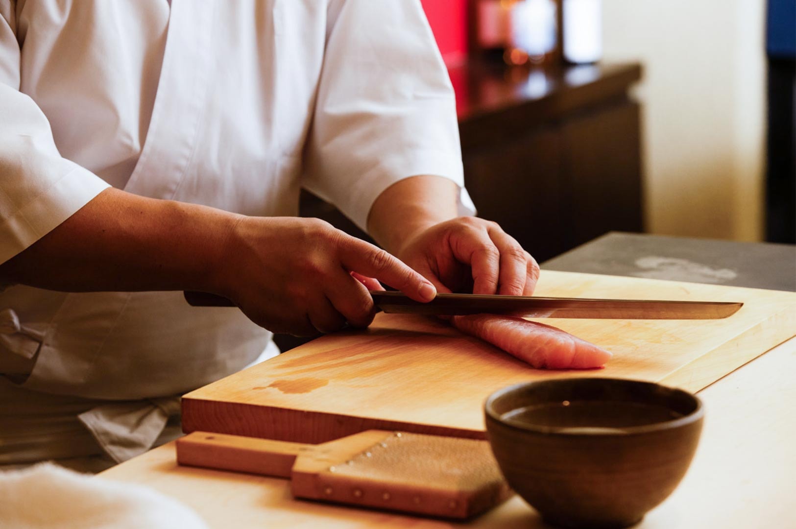 Close-up of a chef’s hands using a traditional Japanese yanagiba knife to precisely slice a fatty piece of tuna on a wooden cutting board.