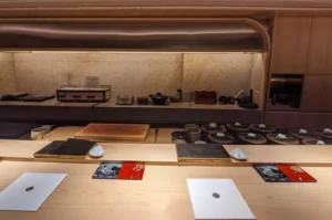 The minimalist interior of Hamamoto, an omakase restaurant. A long, light-colored wooden counter dominates the foreground, with individual place settings featuring black slate plates and small white ceramic rests. In the background, a warm, textured stone wall sits behind a prep area equipped with cooking tools and traditional kitchenware.