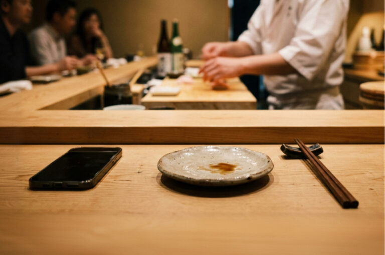 Minimalist hinoki wood sushi counter. In the foreground, an empty ceramic plate with a small soy sauce smudge sits between a pair of wooden chopsticks and a face-down smartphone. The background shows the blurred, motion-filled hands of a sushi chef, capturing a quiet, authentic moment of an omakase experience.