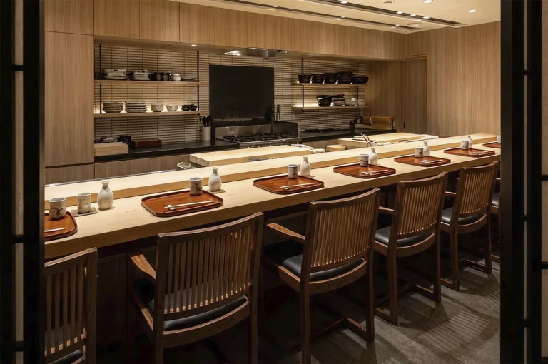 A wide shot of an empty, minimalist Kappo restaurant interior featuring a long, light-colored wooden counter. The counter is set with individual wooden trays, ceramic sake carafes, and cups. In the background, an open kitchen with clean wooden cabinetry and organized shelves of ceramic bowls and plates awaits the chef.