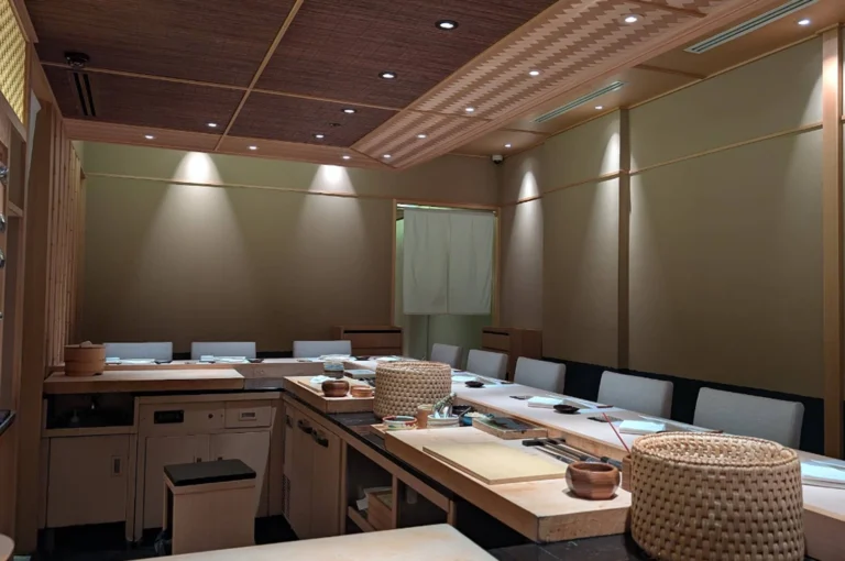 A wide-angle, eye-level shot of a pristine, modern omakase sushi counter. The interior features light-toned wood (likely hinoki) and warm, recessed spotlighting. The L-shaped counter is set with minimalist place settings: white rectangular plates, small dark sauce dishes, and neatly folded white napkins. In the foreground, traditional woven baskets and wooden prep blocks sit ready for the chef. The background shows clean, beige walls and a traditional white noren curtain hanging in a doorway, creating a serene and orderly atmosphere.