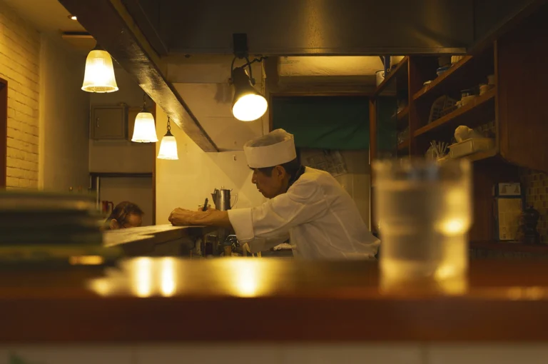 An interior, low-light shot from behind a wooden counter looking toward a sushi chef. The chef, wearing a white uniform and a traditional hat, is focused on writing or studying a document on the counter. The scene is illuminated by warm, hanging pendant lights, creating a quiet, contemplative atmosphere in a Japanese restaurant.